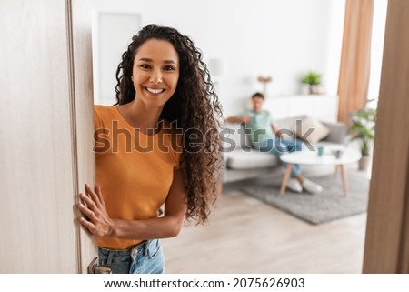 Welcome. Portrait of cheerful lady inviting guests to enter home, happy young lady standing in doorway of modern flat, holding door looking out, man sitting on couch in blurred background Royalty-Free Stock Photo #2075626903