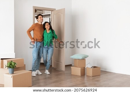 Relocation Concept. Happy millennial newlyweds walking in new empty house with cardboard boxes on floor, young family of two people moving into bought apartment, excited guy and lady choosing flat Royalty-Free Stock Photo #2075626924