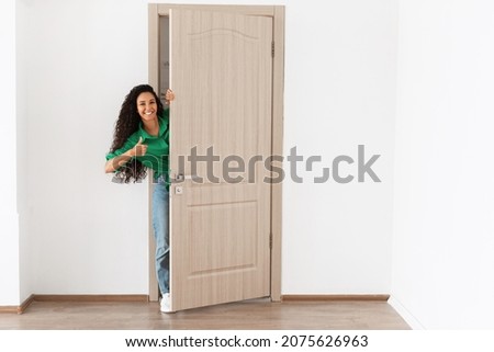 Portrait of cheerful woman standing in doorway of modern apartment, millennial female homeowner holding door looking out and smiling, greeting visitor, showing thumbs up like sign gesture, approving Royalty-Free Stock Photo #2075626963