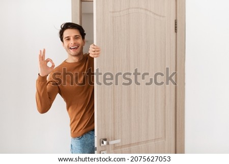 Portrait of cheerful man standing in doorway of modern apartment, millennial male homeowner holding door looking out and smiling, greeting visitor, showing okay sign gesture, approving