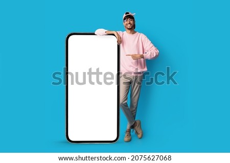 Stylish indian guy standing by huge smartphone with blank screen, mockup, pointing at advertisement, smiling millennial man recommending newest mobile application, blue studio background, copy space Royalty-Free Stock Photo #2075627068