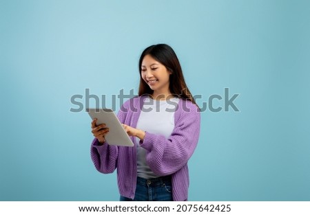 Online life. Asian lady using tablet pc for remote education, standing over blue studio background, communicating on internet or scrolling social networks feed