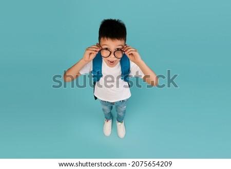 Educational Ad. Top View Shot Of Funny Shocked Little Asian Schoolboy In Glasses, Emotional Korean Male Child Wearing Backpack Looking At Camera With Excitement, Standing Over Blue Studio Background
