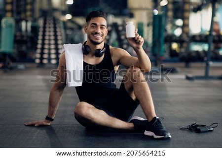 Young Muscular Arab Man Holding Container With Supplement Pills, Middle Eastern Male Athlete Posing At Gym With Blank Pack Of Multivitamins In Hands, Sitting On Floor In Sport Club And Smiling Royalty-Free Stock Photo #2075654215