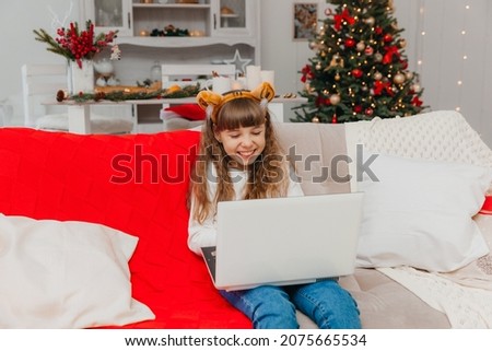 A little girl in a santa claus hat communicates by video call via a laptop, sitting on the couch in the living room.