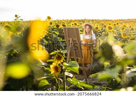 A woman is standing in a field of sunflowers and drawing a picture. The canvas is standing on an easel. She is holding a brush and palette. Sunflowers in the foreground.