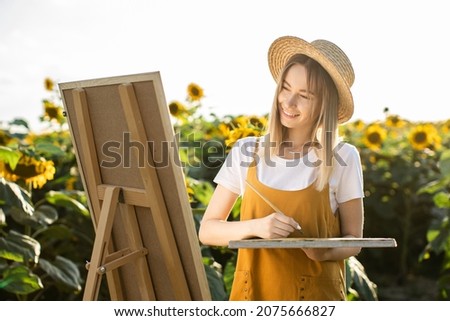 A woman is standing in a field of sunflowers and drawing a picture. The canvas is standing on an easel. She is smiling and looking at the picture. She is holding a brush.