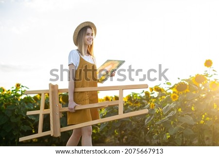 A young woman is carrying an easel and a painting. She is walking through a field of sunflowers.