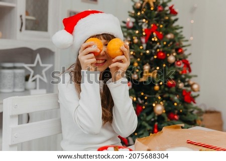 Happy Caucasian girl in a red Santa hat holds tangerines in her hand. There is a Christmas tree in the background.