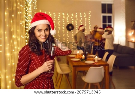 Happy good looking young single woman in polka dot dress and Santa cap holding wine glass and smiling at camera standing against background of decorated interior on Christmas party night with friends