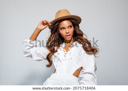 Fashion Portrait Black Woman in white stylish linen dress. Makeup, curly Hair. Luxury Fashion model African American posing in studio against a light wall. Beautiful Black Woman in beige felt Hat      Royalty-Free Stock Photo #2075718406