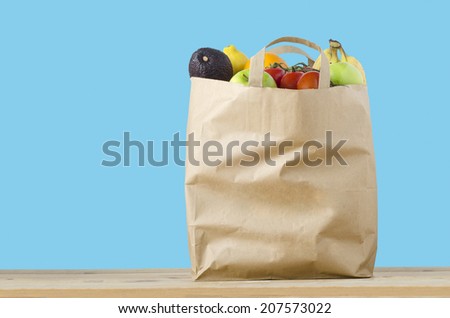 A brown paper shopping bag, filled to the top with varieties of fruit, on a light wood surface.  Isolated on a turquoise blue background. Royalty-Free Stock Photo #207573022