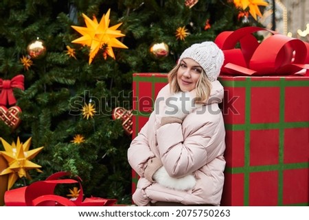 young woman in warm light clothes and white mittens against the background of a huge box with a gift in human growth and an elegant Christmas tree