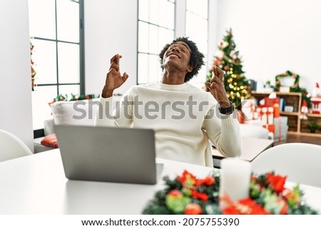 Young african american man using laptop sitting on the table by christmas tree gesturing finger crossed smiling with hope and eyes closed. luck and superstitious concept. 