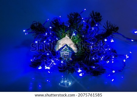 Christmas tree toy in the form of a house lies on a Christmas tree branch under the light of lights