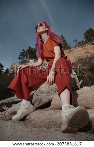 a woman with pink hair in sunglasses and orange clothes sits on a log on the beach and looks at the sun