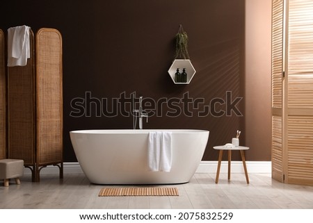 Modern ceramic bathtub with towel near brown wall in room Royalty-Free Stock Photo #2075832529