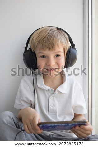 cute boy 4-5 years old in big headphones and with a mobile phone, sits alone, plays games, watches cartoons or listens to music on a smartphone. Digital generation and telephone addiction concept