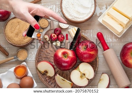 Female hand with mobile phone taking picture ofi for Apple pie or Charlotte on light wooden table. Ingredients for apple pie - red apples, flour, eggs, sugar, cinnamon and butter on a wooden table