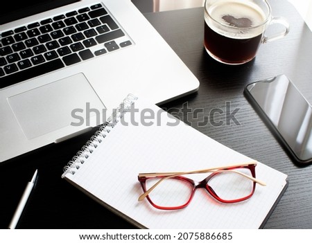 Flatlay on the topic of working at a computer. Laptop, notepad, pen, mobile phone, a cup of coffee and women's glasses in red frames. Items on a black background. High quality photo