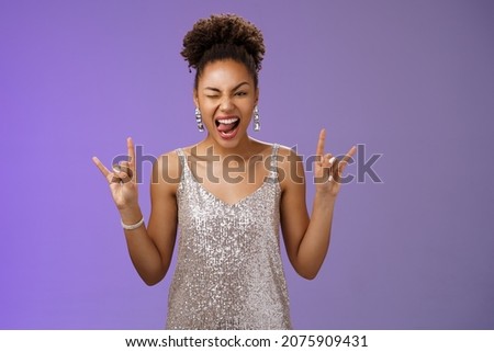 Excited good-looking african-american woman in glittering silver shiny dress having fun amusing university party prom night show tongue winking joyfully rock-n-roll heavy-metal gesture, blue wall