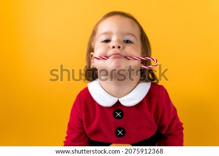 Portraite Cute Cheerful Chubby Baby Girl in Santa Suite Holding Eating Caramel Candy At Yellow Background. Child Play Christmas Scene Celebrating Birthday. Kid Have Fun Spend New Year Time Copy Space