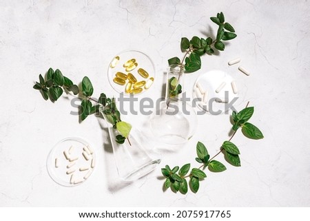 Natural herbal medicine concept. White and yellow medical pills in Petri dishes and laboratory glassware with fresh green plants on white stone background top view. Vitamins and omega oils for vegans.
