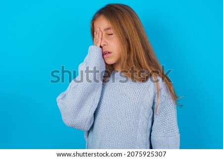brunette kid girl wearing blue knitted sweater over blue background Yawning tired covering half face, eye and mouth with hand. Face hurts in pain.