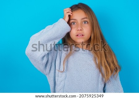 Embarrassed brunette kid girl wearing blue knitted sweater over blue background with shocked expression, expresses great amazement, Puzzled model poses indoor