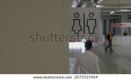 Close up of male and female washroom logo on a beige wall inside a business center. HDR. Back view of a man in white shirt passing by the public toilet.