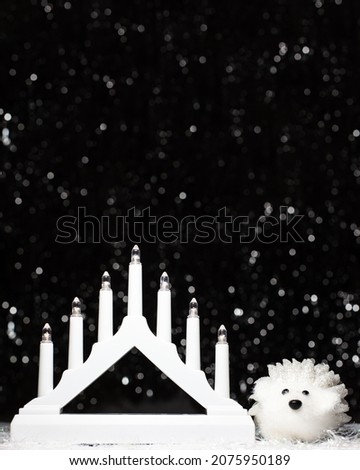 Christmas banner decorations view of white candle lights bridge and white hedgehog on dark background with silver colors bokeh and artificial snow. Holidays concept with copy space at the top
