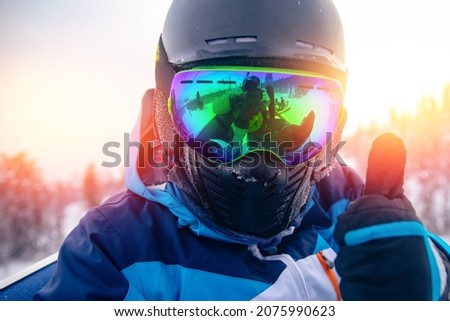 Skier man in balaclava, helmet and glasses is taking selfie in snow on mountain ski lift, sun light.