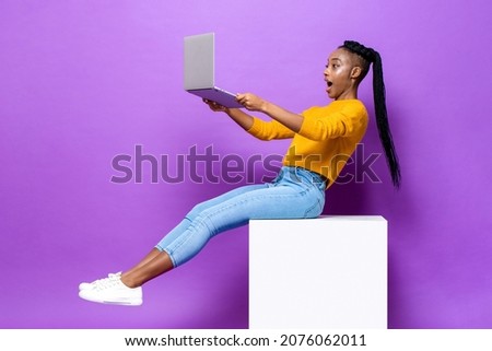 Excited African American woman holding and watching  laptop computer while sitting on stool in isolated purple color studio background Royalty-Free Stock Photo #2076062011