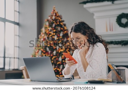 Young happy spanish businesswoman using smartphone while working laptop at home on Christmas, sitting at desk against fir xmas tree with lights, reading sms message on mobile during winter holidays Royalty-Free Stock Photo #2076108124