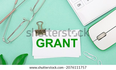 On a light green background there is a white calculator, a computer mouse, green leaves of a plant, gold-rimmed glasses and a white card with text GRANT. Business concept