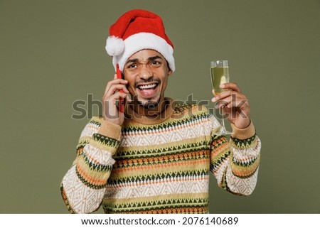 Young african man in sweater red Christmas Santa Claus hat speak talk mobile cell phone hold champagne wineglass greet isolated on plain green khaki background Happy New Year 2022 celebration concept