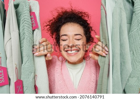 Close up young fun smiling excited happy cheerful female costumer woman 20s wearing sweater stand near clothes rack with tag sale in store showroom isolated on plain pink background studio portrait.