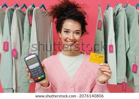 Young smiling woman in sweater stand near clothes rack with tag sale in store showroom hold wireless bank payment terminal process acquire credit card payments isolated on plain pink background studio