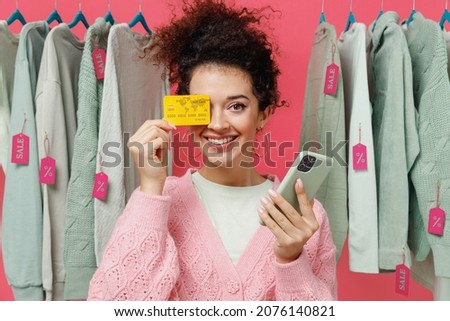 Young smiling woman in sweater stand near clothes rack with tag sale in store showroom using mobile cell phone cover eye with credit bank card shopping online isolated on plain pink background studio
