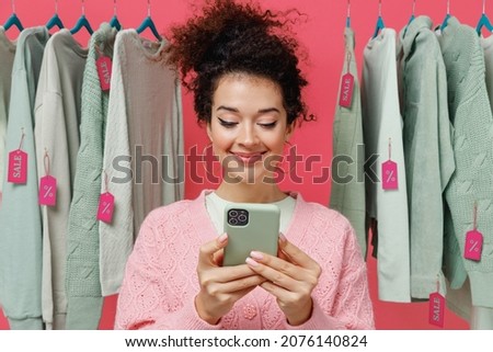 Young smiling happy female costumer woman 20s wear sweater stand near clothes rack with tag sale in store showroom hold in hand use mobile cell phone isolated on plain pink background studio portrait
