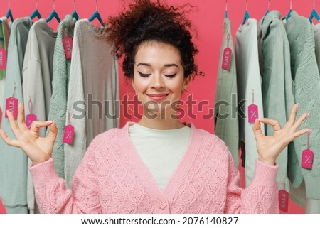Young costumer woman in sweater stand near clothes rack with tag sale in store showroom hold spreading hands in yoga om gesture relax meditate try to calm down isolated on plain pink background studio