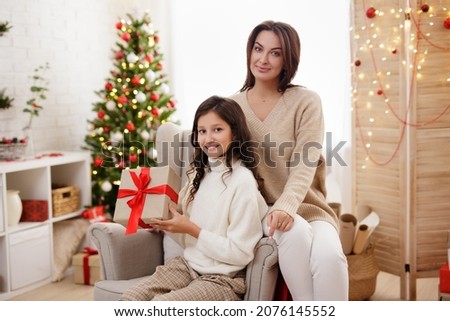 Family and Christmas concept - portrait of happy mother and cute daughter sitting in decorated living room with Christmas tree and christmas gifts