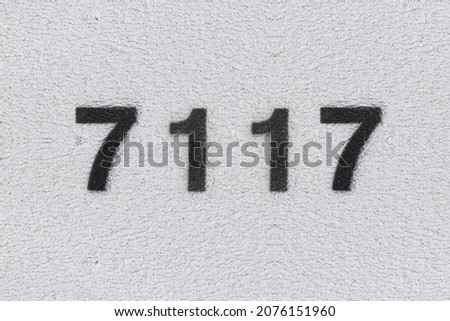 Black Number 7117 on the white wall. Spray paint. Number seven thousand one hundred and seventeen.