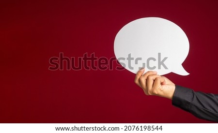 A speech bubble concept. Hand holding an empty white speech bubble over on a red background with copy space for text. Close-up photo.
