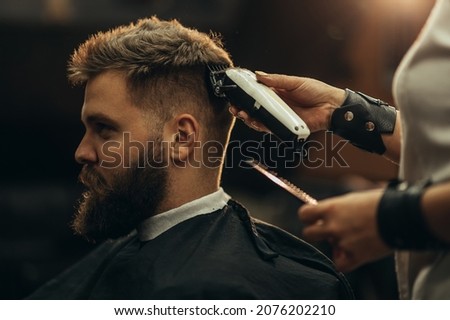 Young bearded man getting haircut by hairdresser while sitting in chair at barbershop Royalty-Free Stock Photo #2076202210
