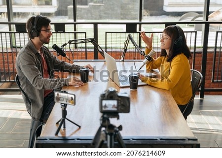 Two young bloggers, man and woman in headphones and glasses looking at each other while talking, recording conversation, interview for video blog. Female host interviewing guest for her video podcast