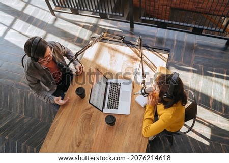 Top view of two people in headphones recording audio podcast using microphones in broadcast studio. Young female talking with her male colleague while they recording podcast. Live streaming