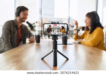 Man and woman hosts talking with each other while recording live video podcast in studio. Two podcasters in headphones shooting live video using smartphone on tripod. Selective focus on smartphone