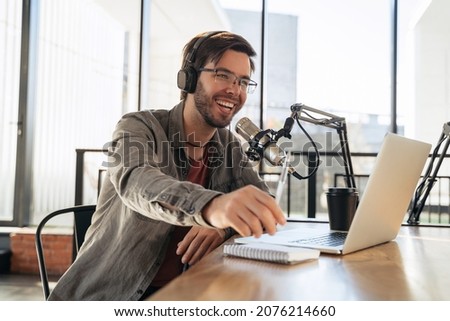 Young man host in headphones and glasses enjoying podcasting in studio, speaking into a microphone, holding a pen, using laptop. Handsome podcaster laughing while streaming live audio podcast Royalty-Free Stock Photo #2076214660