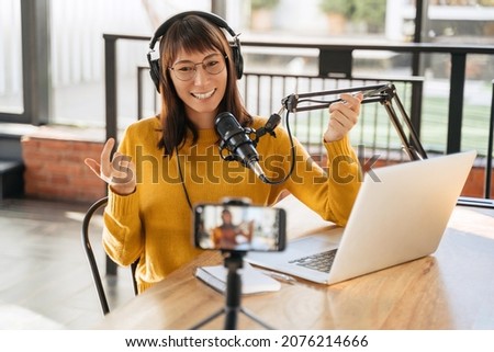 Woman podcaster in headphones and glasses looking at smartphone camera, gesticulating, laughing, using microphone, shooting video podcast on laptop in studio. Female podcast creator recording podcast